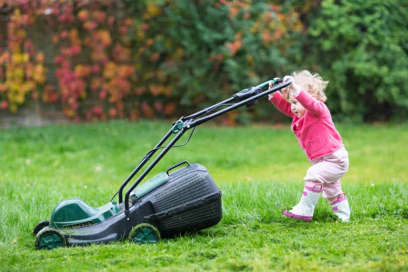 Cute curly baby girl in rain boots playing in a big green lawn with walk behind lawn mower
