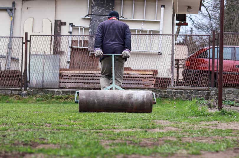 Adult man make garden smoother with help lawn roller. Summer volunteer pulls heavy roller behind and aligns the surface.